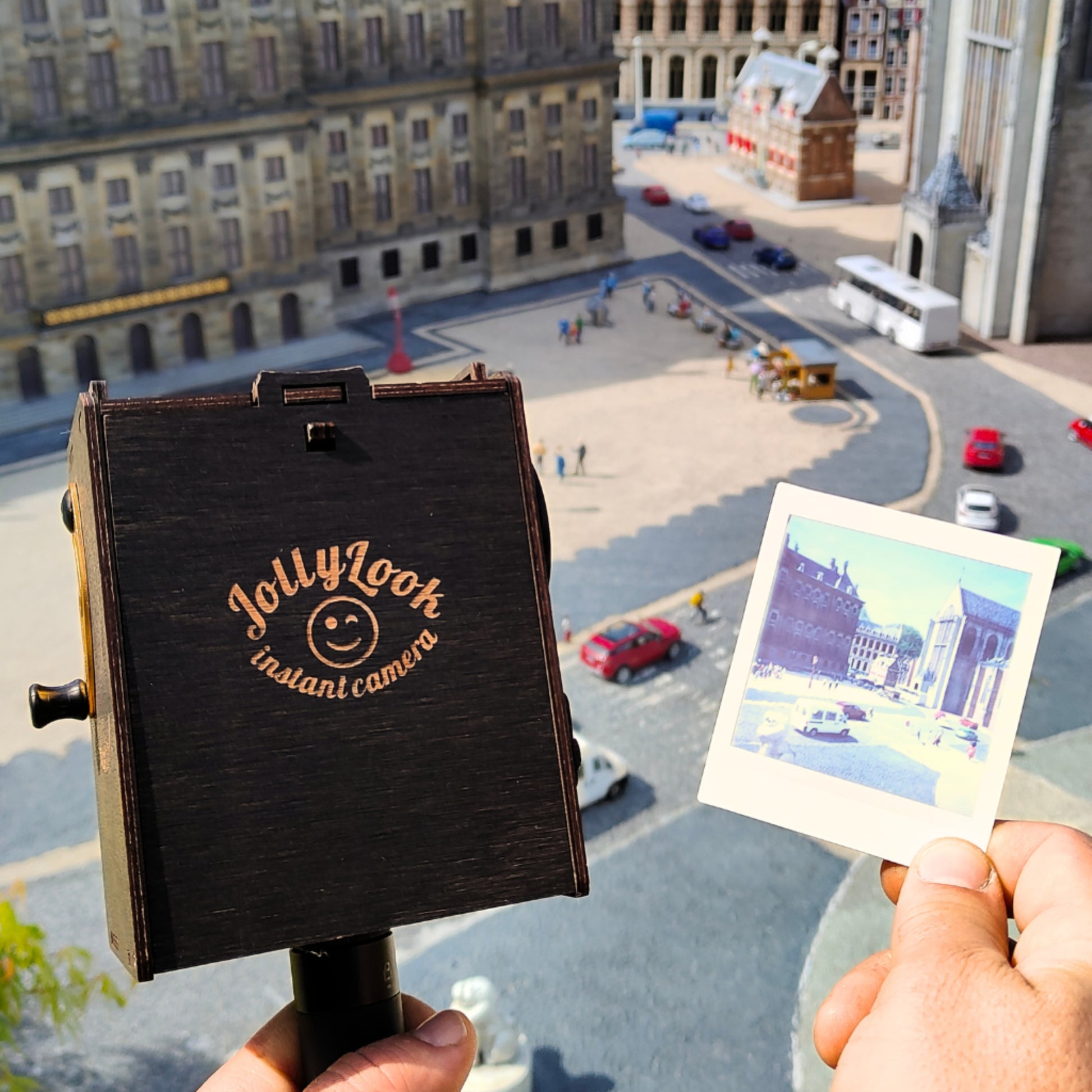 A moment where a Pinhole SQUARE Instant Film Camera in Stained Brown color is held in the left hand, its vintage aesthetic complementing the timeless appeal of an ancient city unfurling in the backdrop. In the right hand, a freshly snapped instant photo of a city scene is displayed, providing a tangible, miniature reflection of the urban tapestry behind.