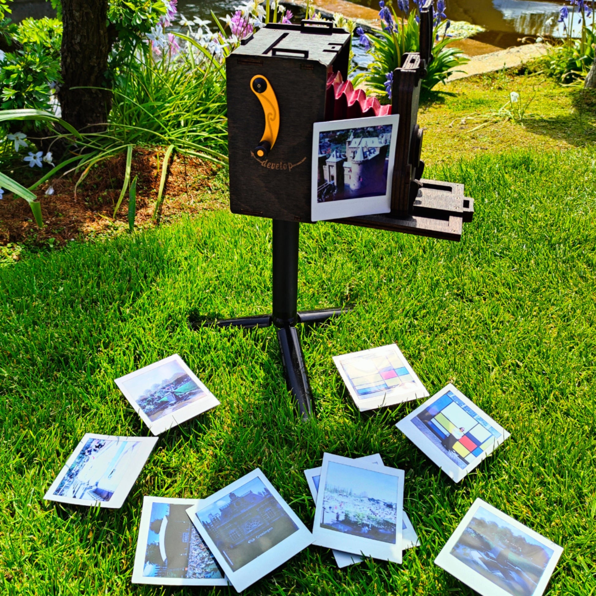 Pinhole SQUARE Instant Film Camera in Stained Brown color perched securely on a tripod, positioned on a vibrant stretch of green grass. Surrounding the camera, a scattering of instant SQUARE photos, each capturing varied scenes, lay whimsically on the grass, creating a juxtaposed narrative of photographed moments in the tangible present.