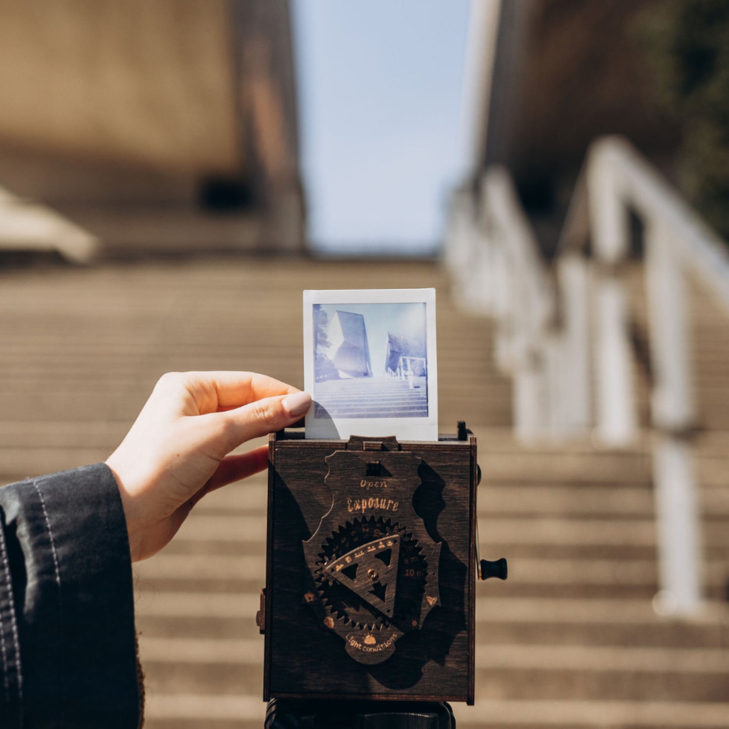 Picture showcasing the back of the Pinhole SQUARE Instant Film Camera in Stained Brown Color juxtaposed against an urban staircase backdrop within a cityscape. In a complementary layer, a hand elegantly holds a freshly captured photo, taken with the camera, depicting the same staircase.
