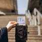 Picture showcasing the back of the Pinhole SQUARE Instant Film Camera in Stained Brown Color juxtaposed against an urban staircase backdrop within a cityscape. In a complementary layer, a hand elegantly holds a freshly captured photo, taken with the camera, depicting the same staircase.